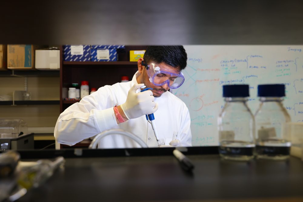 A UNC Charlotte student in the Ren Lab in the Klein College of Science wears a lab coat, gloves and safety glasses while using a pipette.