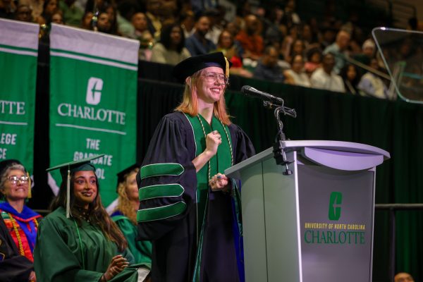 Hal West-Page speaks at the podium on stage during UNC Charlotte's August 2026 commencement ceremony. West-Page wears her Niner green and black doctoral gown and hood for earning a Ph.D. in applied mathematics.