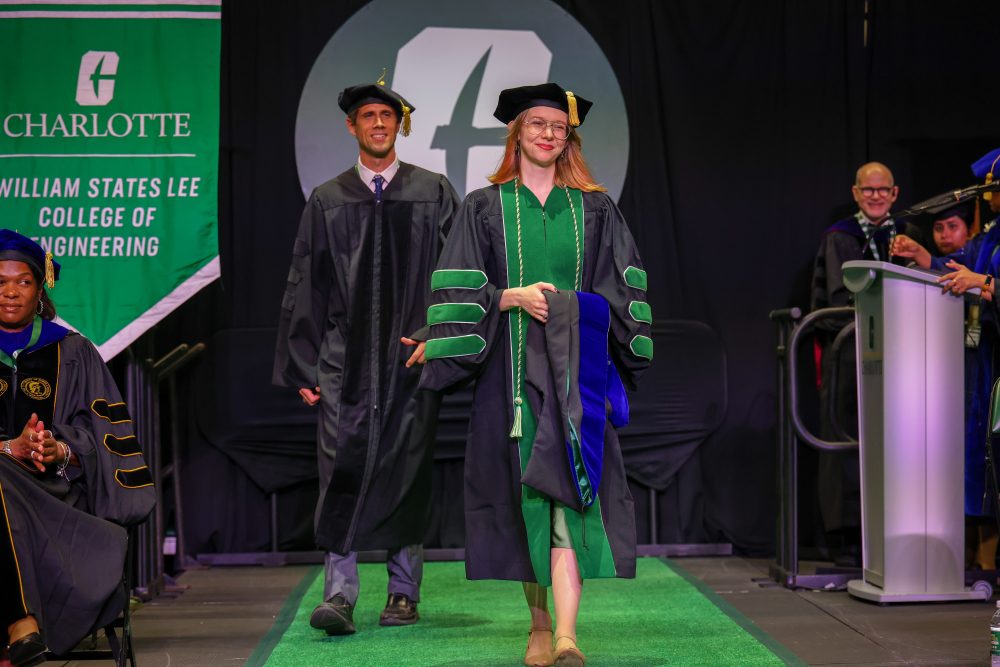 Hal West-Page, three time UNC Charlotte graduate, walks onto the stage during the August 2026 commencement ceremony with her hood draped over her arm for the hooding of her Ph.D. in applied mathematics from the UNC Charlotte Klein College of Science.