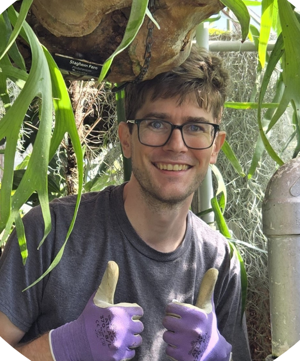 Josh Munn giving two thumbs up to the camera while working in the greenhouse, part of the UNC Charlotte Botanical Gardens