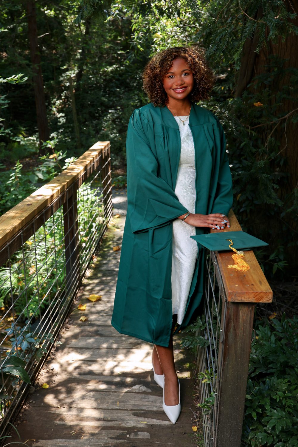 Zarah Rahman Blythe, biology major in the UNC Charlotte Klein College of Science, wears her Niner green graduation gown and stands on a bridge in UNC Charlotte's Botanical Gardens.