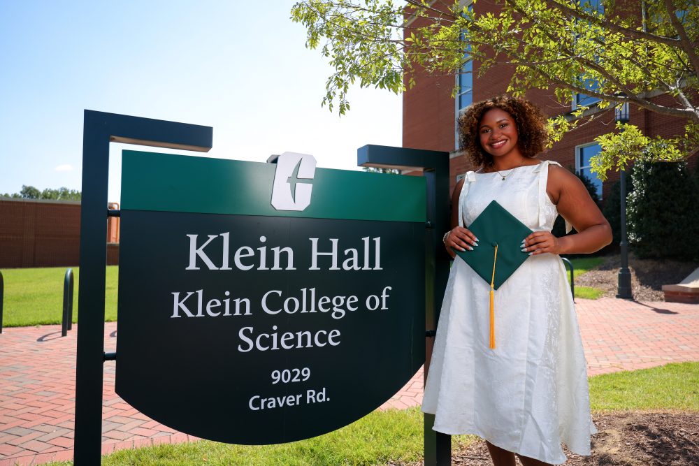 Zarah Rahman Blythe, biology major in the UNC Charlotte Klein College of Science, holds her graduation cap, posed next to the Klein Hall sign on UNC Charlotte's campus.