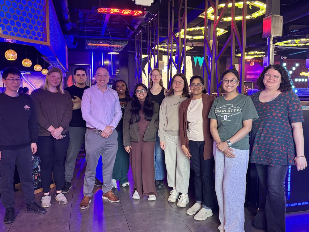 A group of eleven members of the Truman Lab stand smiling inside KPOT Korean BBQ & Hot Pot, a brightly lit restaurant with neon lights and geometric decor, including a sign reading “KPOT.” One person wears a green Charlotte T-shirt.