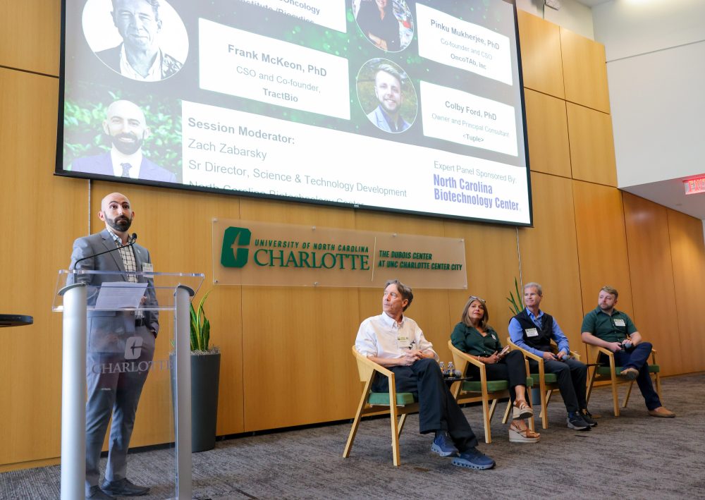 A speaker stands at a podium addressing an audience, with four panelists seated in front of a large screen displaying speaker information and logos for the University of North Carolina Charlotte and the North Carolina Biotechnology Center.
