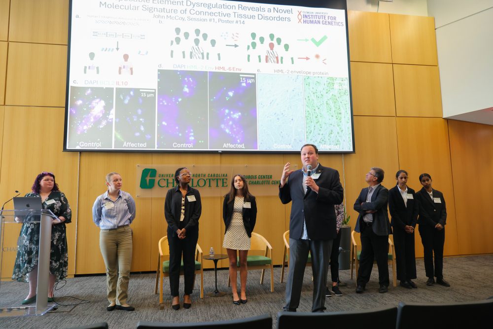 A group of researchers stands on a stage during a scientific presentation, with one person speaking into a microphone while others listen, in front of a large screen displaying a research poster.