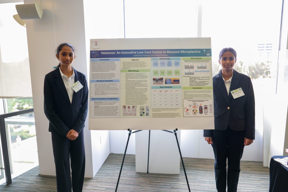 Two young individuals stand beside a research poster titled "Hemovue: An Innovative Low-Cost Device to Measure Microplastics" at a presentation event.