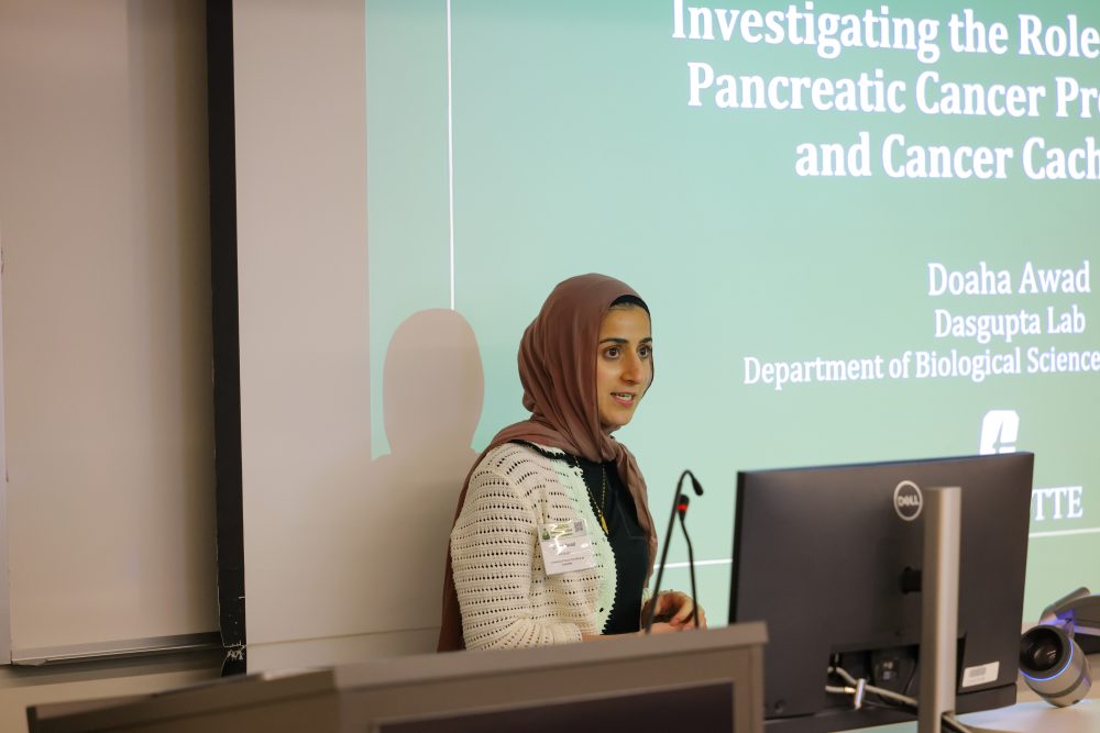 Doaha Award presents at the 2026 Biomedical Sciences Symposium, standing before a screen displaying a talk titled "Investigating the Role of Pancreatic Cancer..." and their affiliation with the Dasgupta Lab.