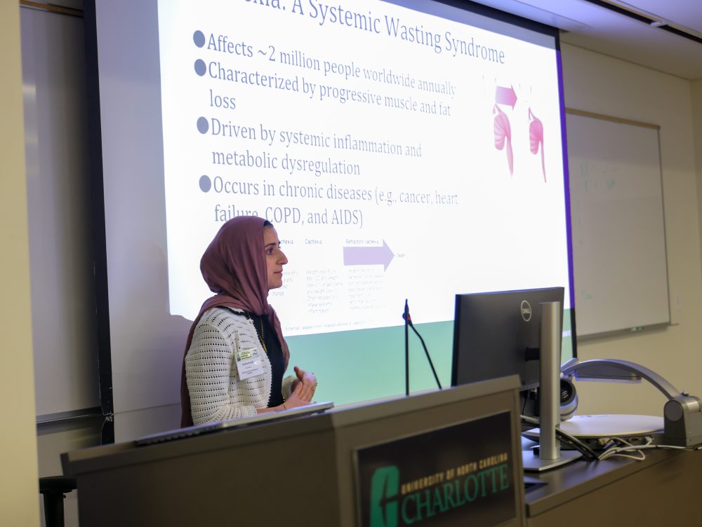UNC Charlotte Dasgupta Lab member Doaha Awad presents in front of a lecture hall at the Biomedical Sciences Symposium, standing beside a screen displaying a slide about the systemic wasting syndrome.