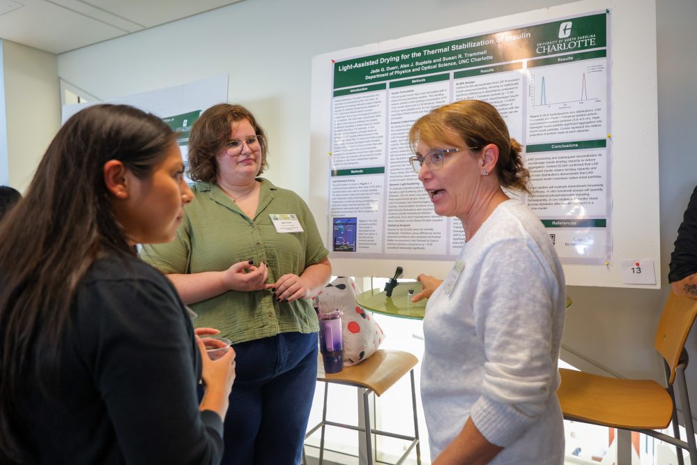 Three individuals engage in conversation in front of a scientific research poster displayed at the University of North Carolina Charlotte.