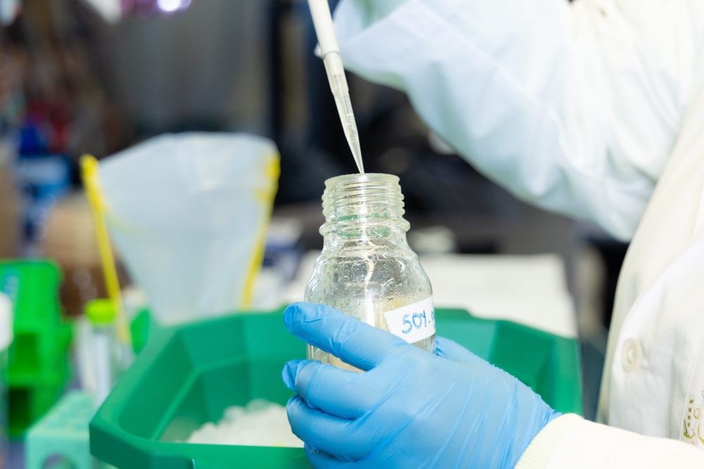 A Truman Lab researcher wearing a white lab coat and blue gloves uses a pipette to transfer liquid into a labeled glass bottle resting in a green ice container.