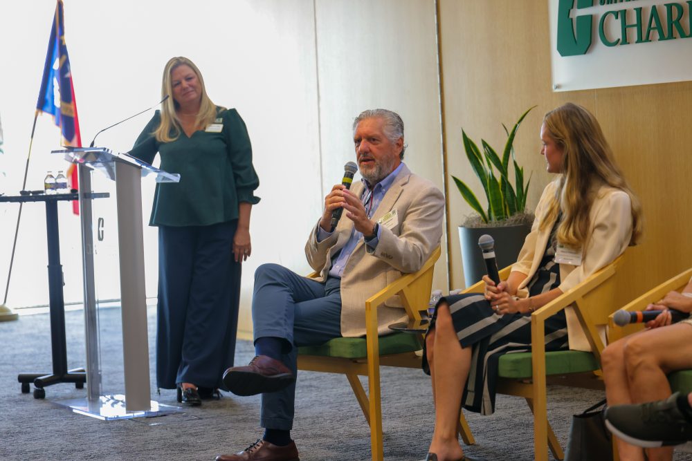 Panelists sit and hold microphones while moderator Christine Richardson stands near the podium at the UNC Charlotte Dubois Center.