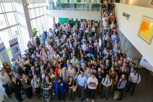 The large crowd of 420+ attendees gather at the 2026 Biomedical Sciences Symposium hosted at the UNC Charlotte Dubois Center on Sept. 14. They stand in the atrium smiling and looking up. Vice Chancellor for Research John Daniels stands at the back of the crowd holding up the pickaxe hand sign.
