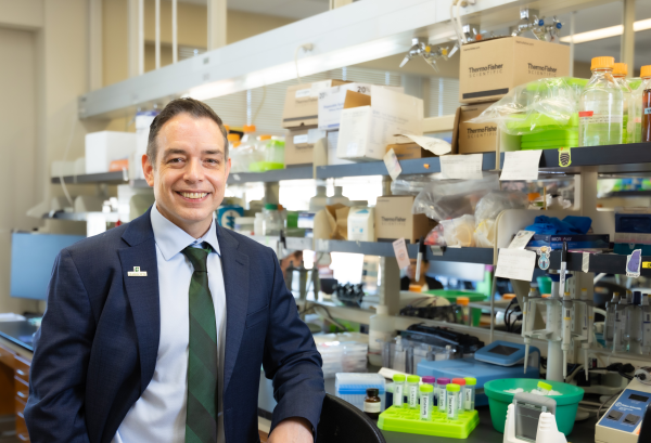 Andrew Truman, associate chair for research in the Klein College of Science's Department of Biological Sciences at UNC Charlotte, wears a dark suit and green tie and smiles while leaning against the counter of his lab filled with scientific equipment including test tubes and bottles.