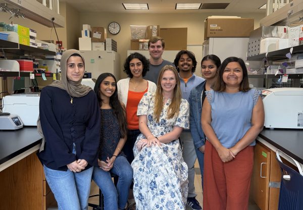 Eight members of the Dasgupta Lab pose together in a brightly lit UNC Charlotte laboratory, standing and sitting between workbenches lined with scientific equipment, boxes and supplies.