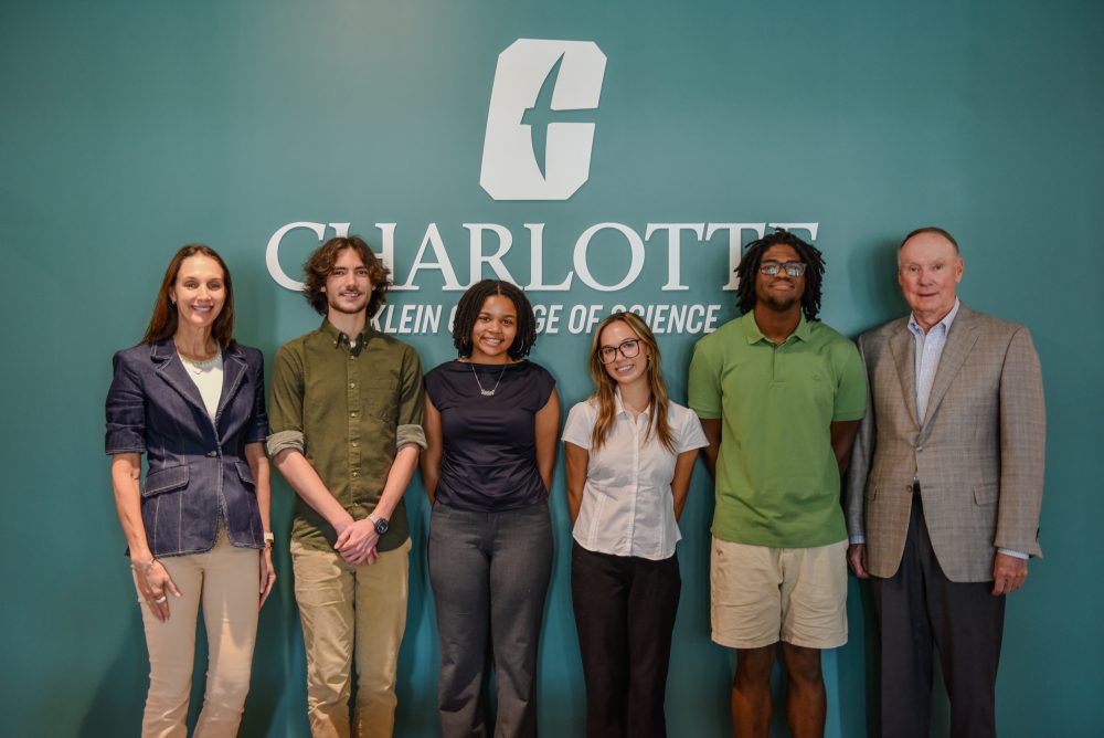 Four UNC Charlotte students selected at Klein Scholars stand in front of a Niner green wall with the KCOS logo, with Fred Klein and Amy Klein Aznar in the lobby of Klein Hall.