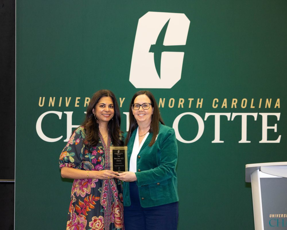 Sejal Foxx and Tonya Bates stand together in front of a University of North Carolina Charlotte backdrop, jointly holding an award plaque.